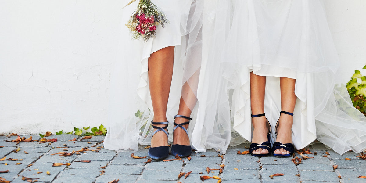 Dos mujeres con zapatos de fiesta azul marino sobre un camino de piedra y vestidos largos blancos
